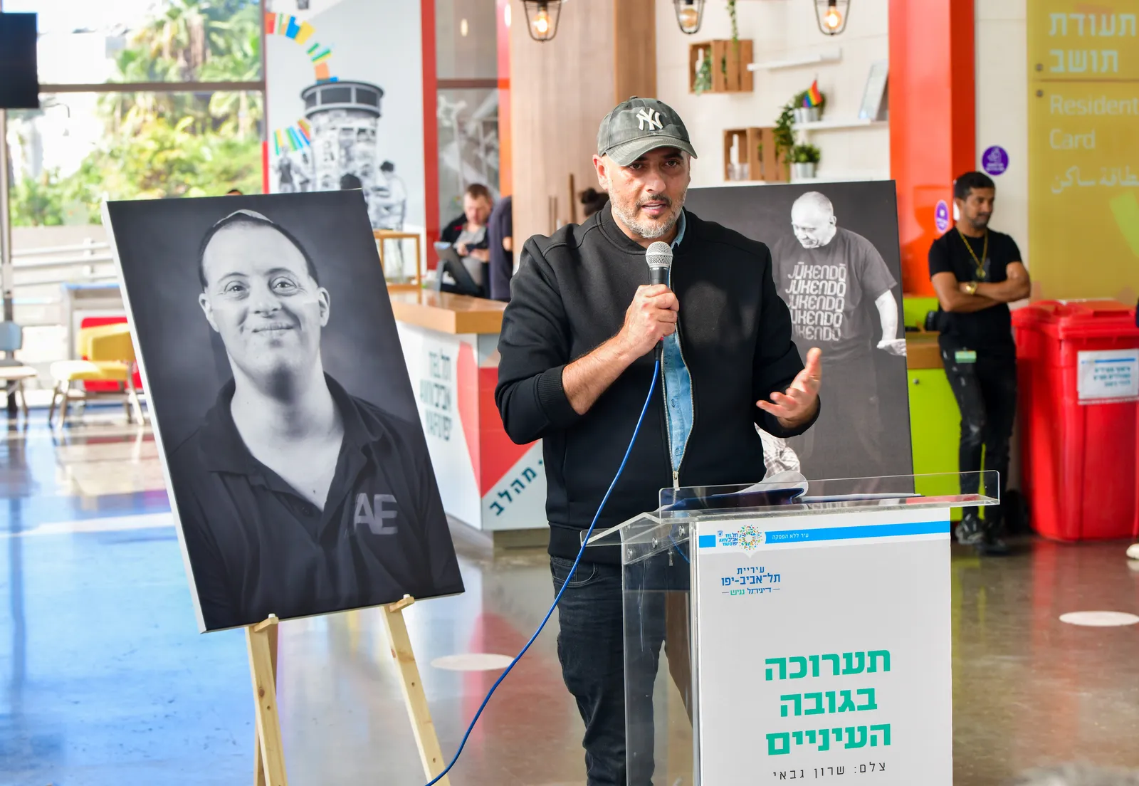 הצלם שרון גבאי נואם בטקס פתיחת התערוכה, לצד פורטרט מהסדרה על כן תצוגה