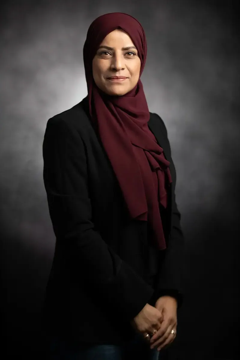 A headshot of a professional in a burgundy hijab and black blazer on a grey backdrop — three-quarter stance, studio lighting
