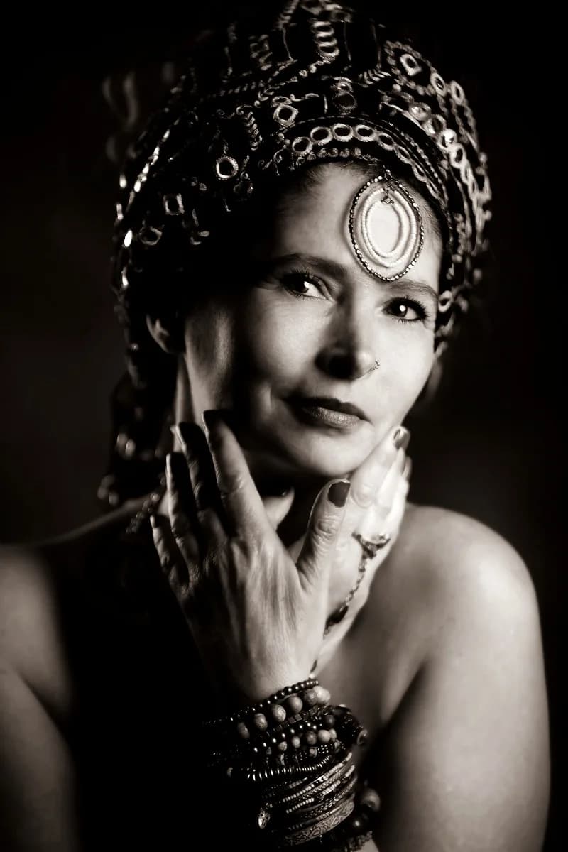 A black-and-white fine-art portrait of a woman in an ornate jewelled headdress, hands to her face — dramatic lighting on a black backdrop