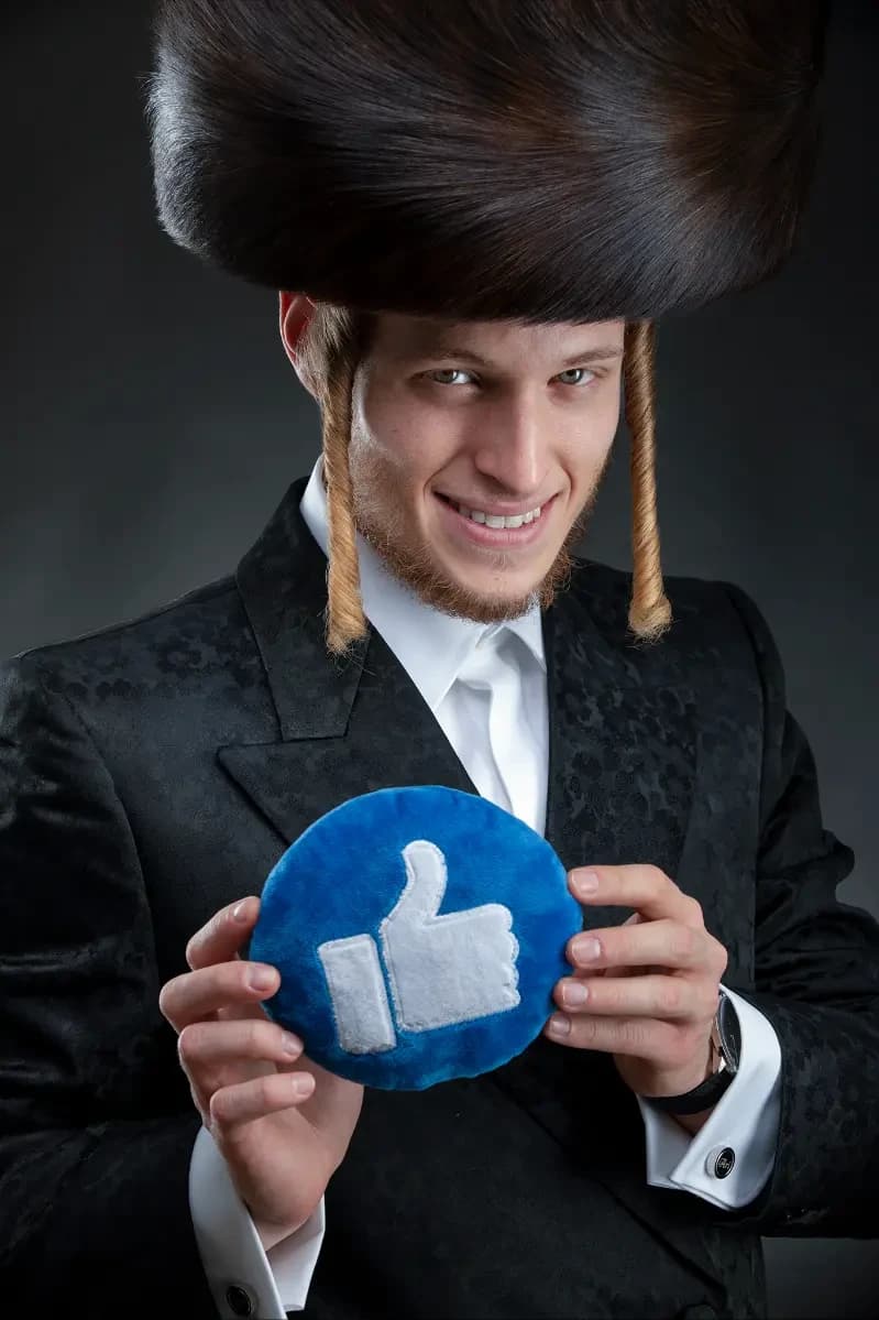 A headshot with a prop: a man in a traditional fur hat and black suit holding a cushion shaped like a “like” icon — a conceptual studio portrait on a dark backdrop