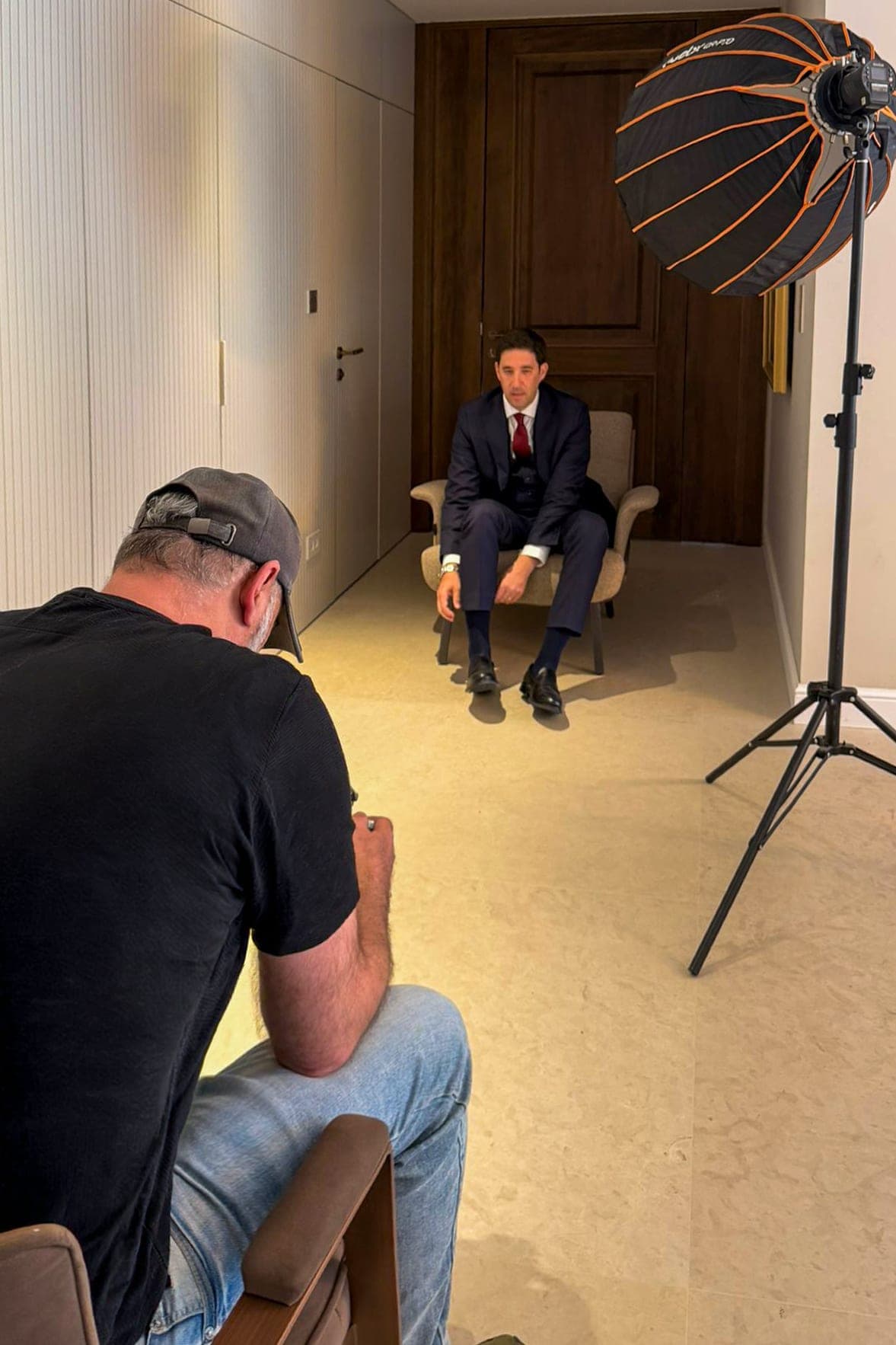 Sharon Gabay seated, photographing a client in a suit seated in an armchair, with studio lighting and a portable backdrop at the client's business