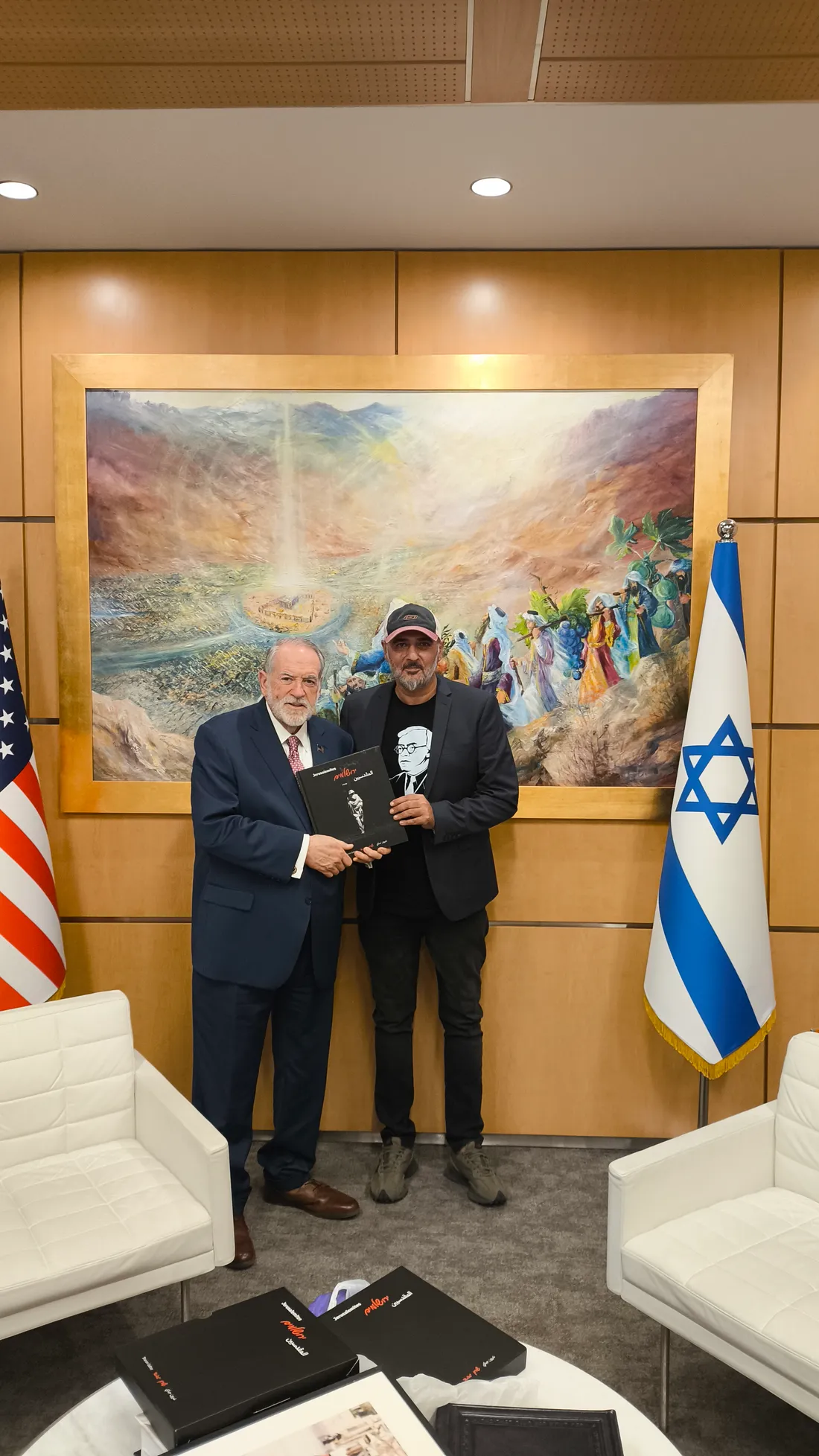 שרון גבאי עם שגריר ארה״ב מייק האקבי, אוחזים יחד את ספר הצילום ״ירושלמים״ על רקע דגלי ארה״ב וישראל