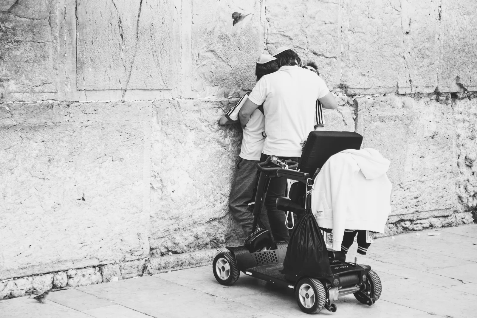 A father lifts his son to kiss the stones of the Western Wall during a bar mitzvah, a bird hovering above — street photograph by Sharon Gabay from the book Jerusalemites