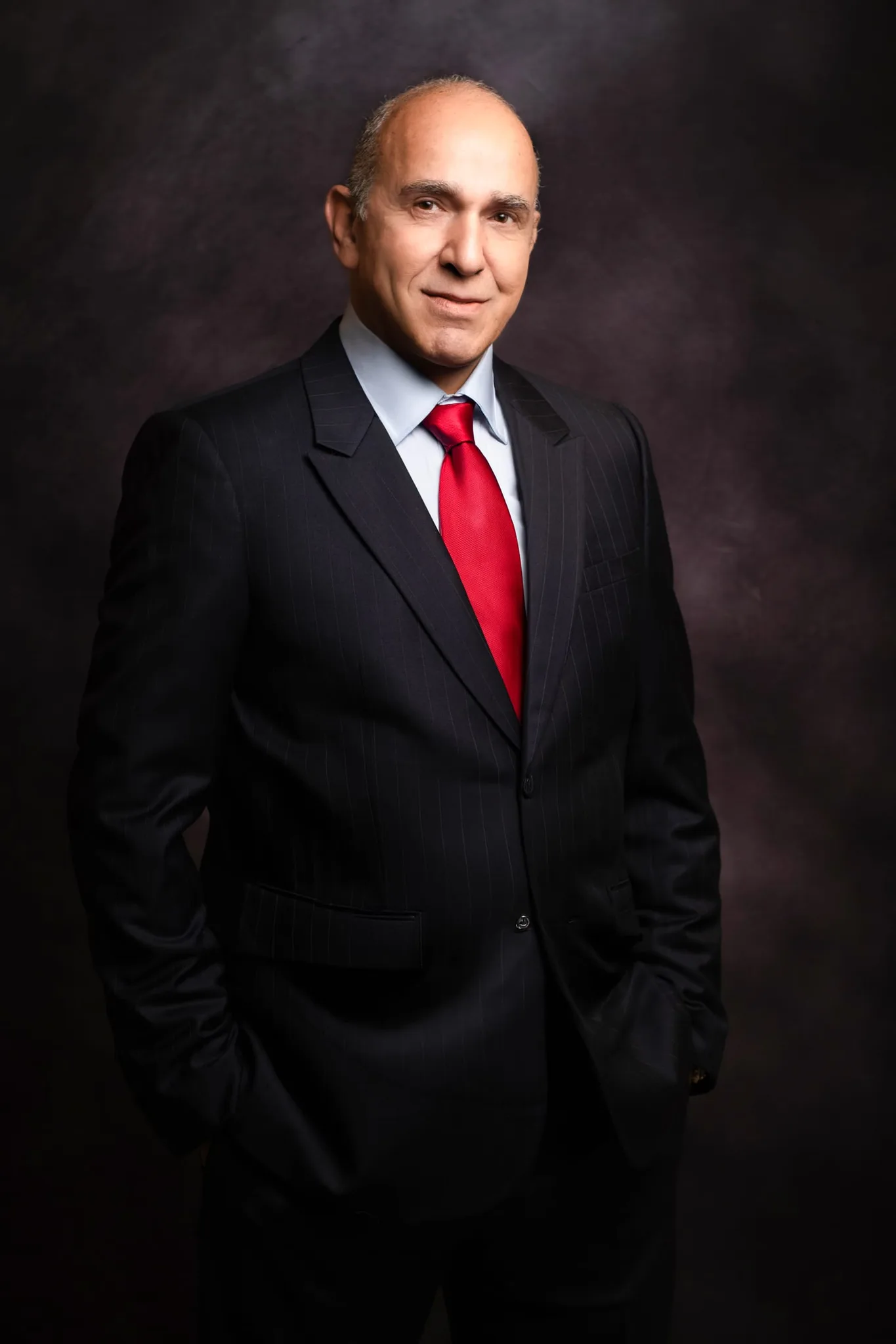 Professional headshot lit with selective light — a man in a dark suit and red tie against a dark background — Sharon Gabay