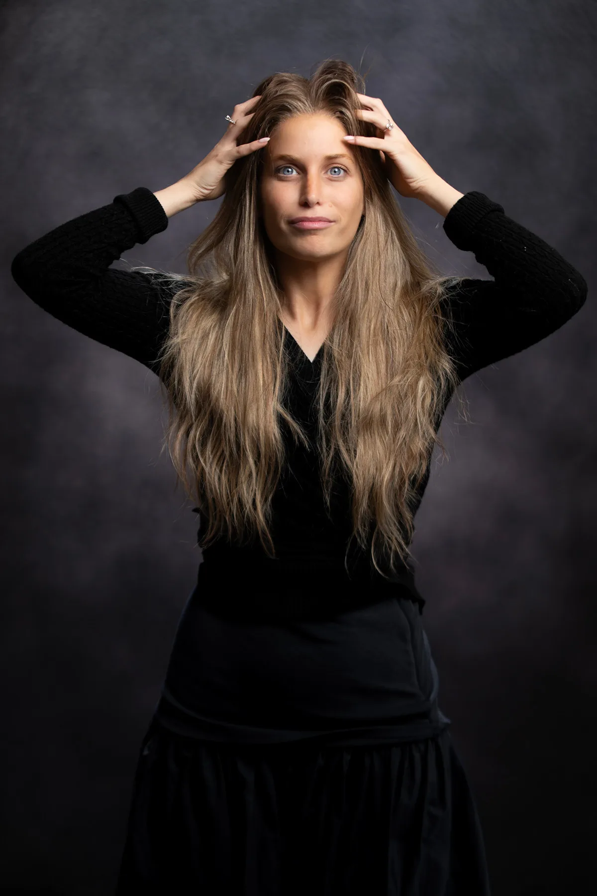 Headshot of a woman in black with long hair, hands in her hair, dramatic studio light | Photo: Sharon Gabay