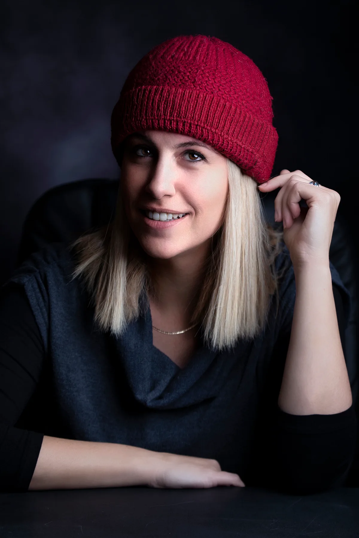 Headshot of a woman in a red beanie leaning on a desk, open smile, soft studio light | Photo: Sharon Gabay
