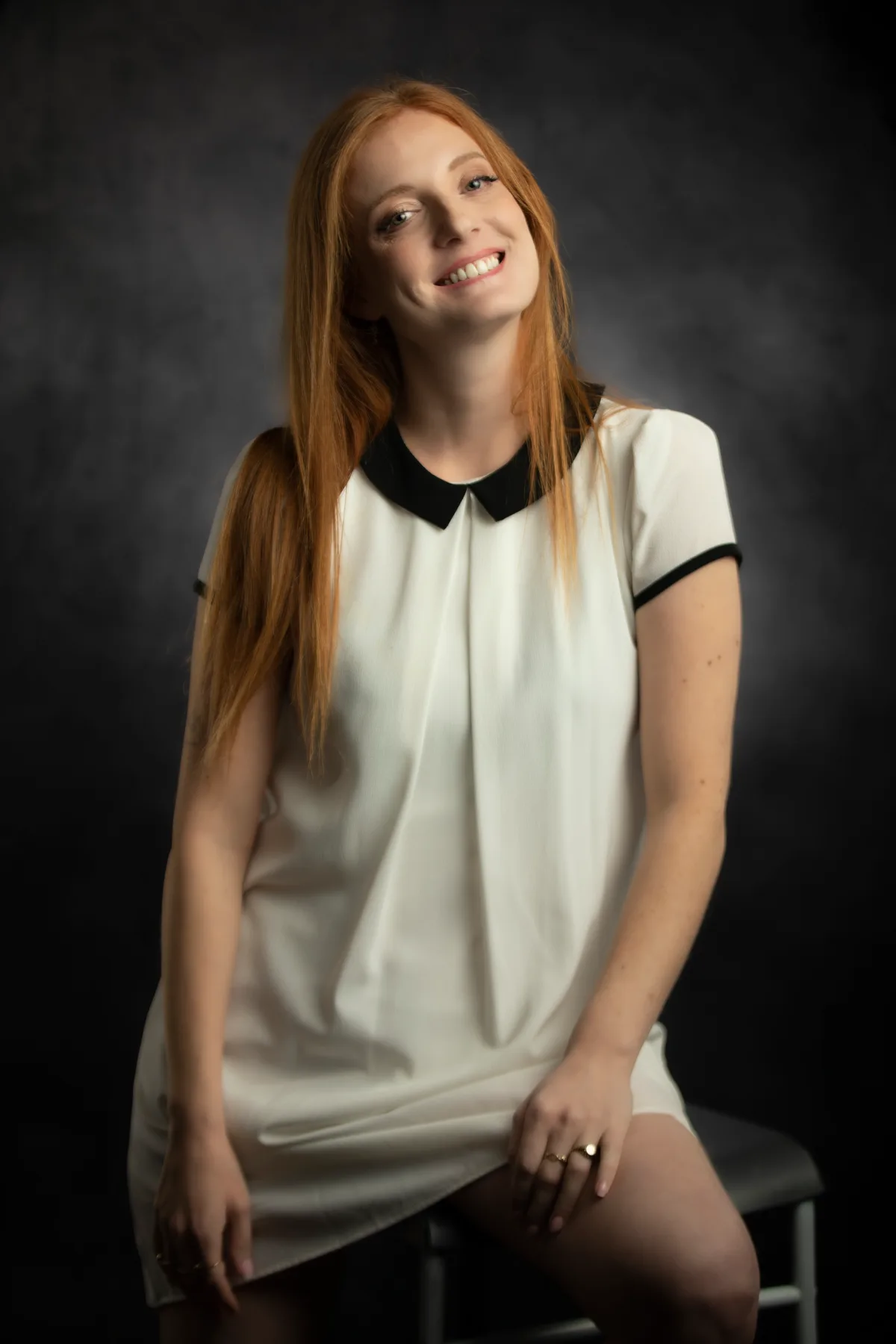 Personal portrait of a red-haired woman in a white dress seated in the studio, soft smile and warm light | Photo: Sharon Gabay